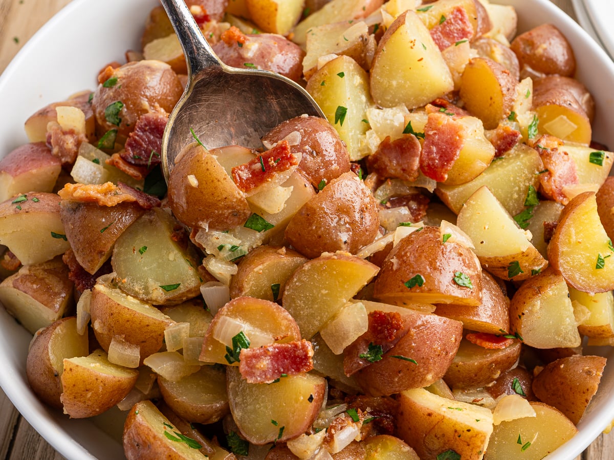 A close-up of a bowl filled with German Potato Salad, made of red potatoes, chopped bacon, onions, and herbs, with a spoon lifting a portion&mdash;perfect for showcasing your favorite German Potato Salad recipe.