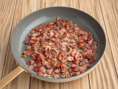 A frying pan with small pieces of crispy, cooked bacon—perfect for a German Potato Salad Recipe—sits on a light wooden surface. The bacon is browned and rendered, with some fat visible in the pan.