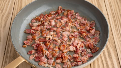 A frying pan with small pieces of crispy, cooked bacon—perfect for a German Potato Salad Recipe—sits on a light wooden surface. The bacon is browned and rendered, with some fat visible in the pan.