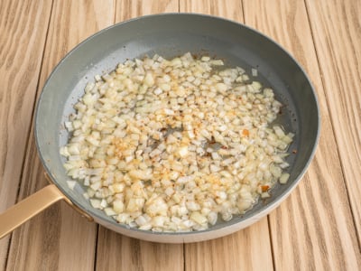 Diced onions sautéing in oil with spices for a classic German Potato Salad recipe in a frying pan on a wooden surface.
