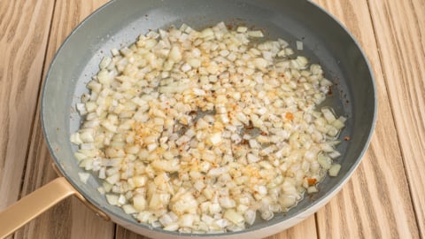 Diced onions sautéing in oil with spices for a classic German Potato Salad recipe in a frying pan on a wooden surface.