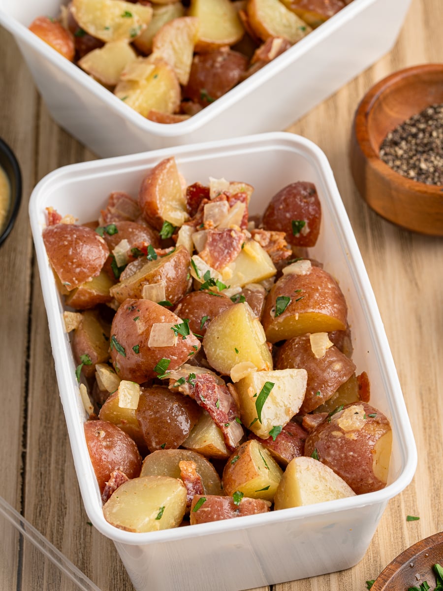A white container filled with chopped red potatoes mixed with bacon, onions, and herbs sits on a wooden surface—an inviting take on a classic German Potato Salad Recipe. Another similar container and a small bowl of pepper appear in the background.