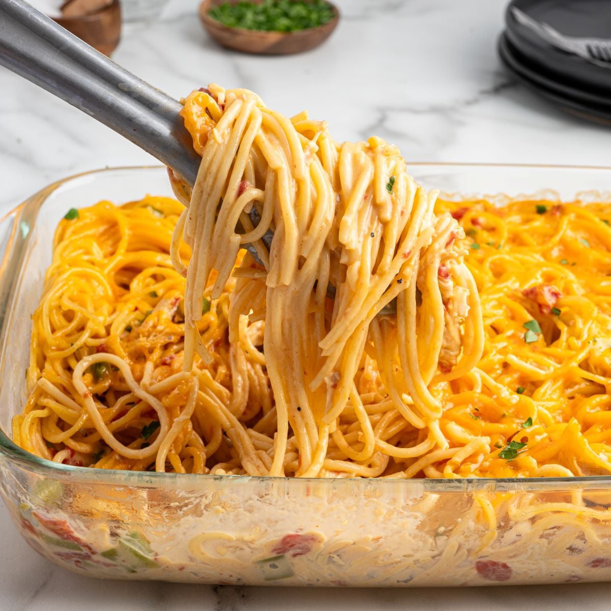 A close-up of tongs lifting a serving of creamy Rotel Chicken Spaghetti from a glass casserole dish, with melted cheese and herbs visible on the pasta.