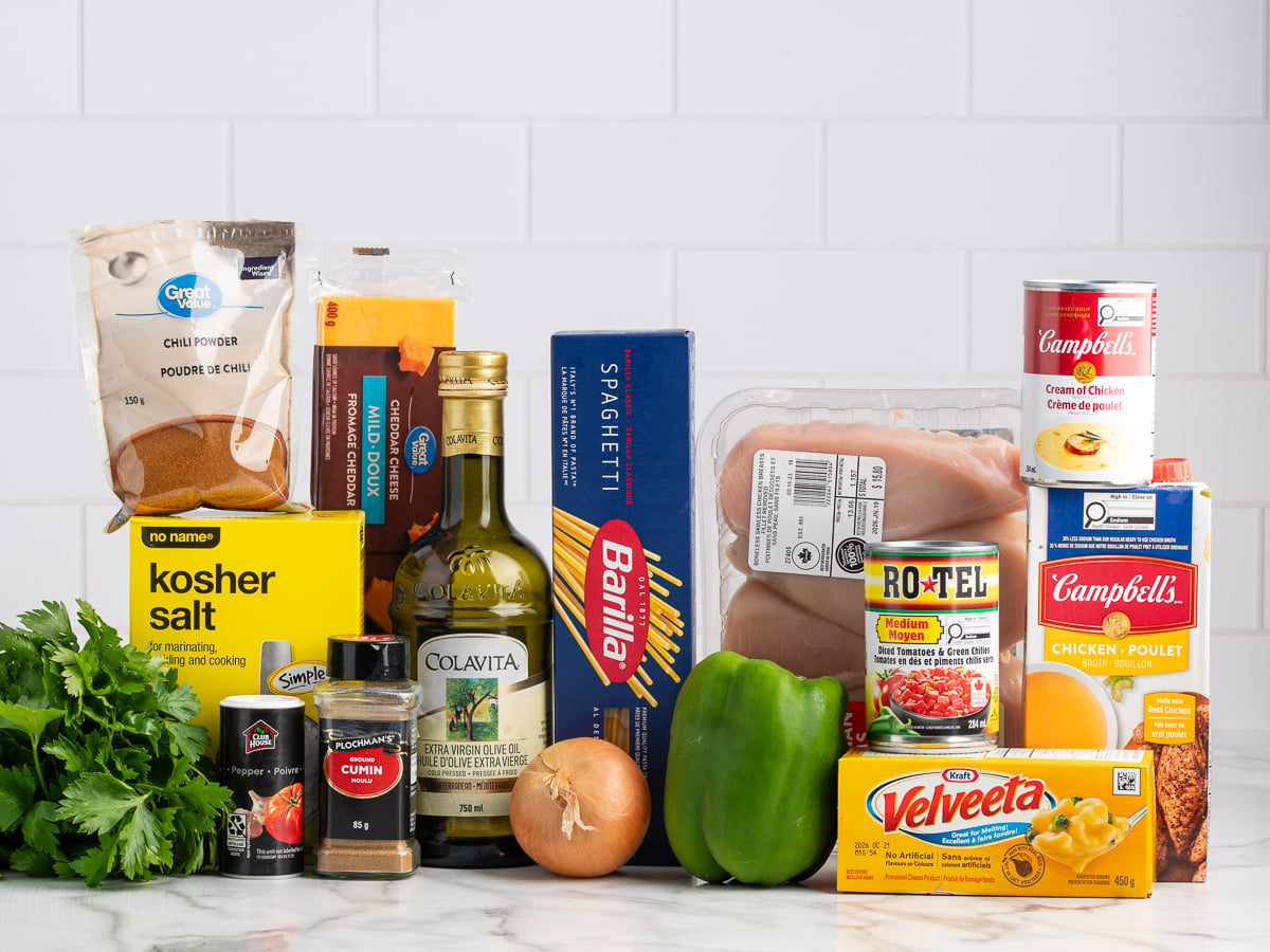 A variety of ingredients for Rotel Chicken Spaghetti—including Velveeta cheese, spaghetti, canned soups, Rotel tomatoes, chicken breasts, green bell pepper, onion, cilantro, olive oil, spices, and kosher salt—are arranged on a counter against a white tile background.