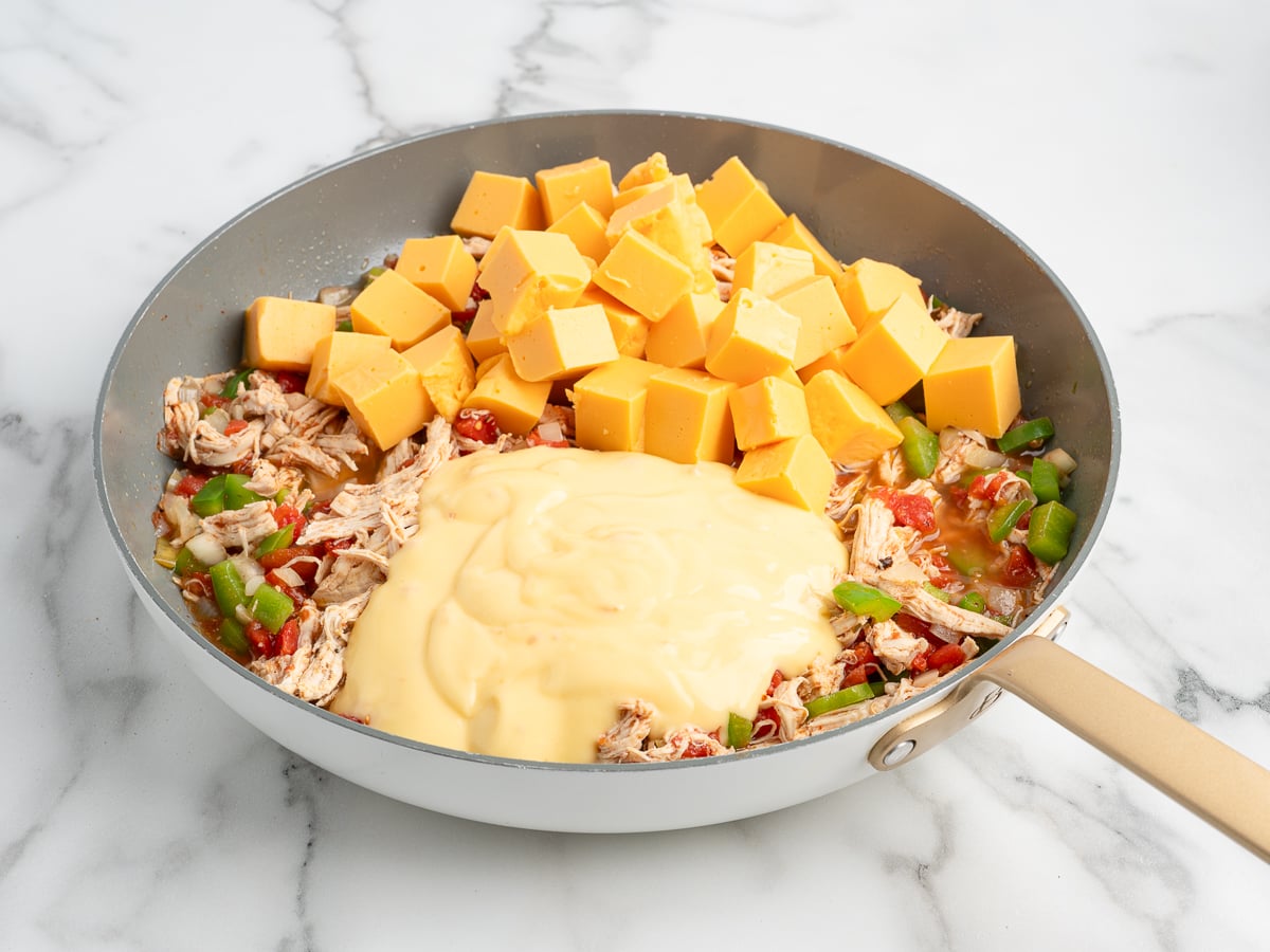 A skillet containing shredded chicken, diced green bell peppers, tomatoes, cubed cheddar cheese, and a creamy cheese sauce layer—this Rotel Chicken Spaghetti-inspired dish sits invitingly on a marble countertop.