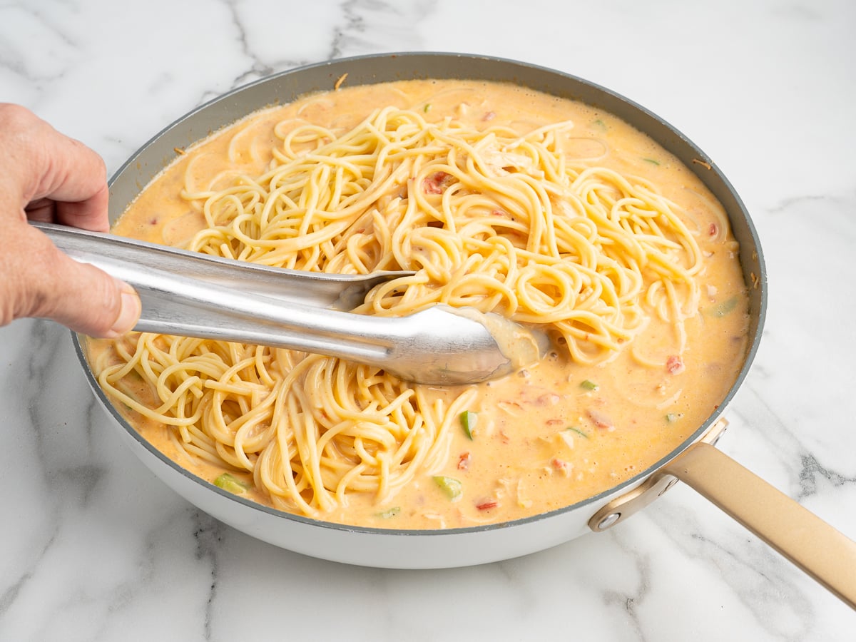 A hand uses metal tongs to mix cooked Rotel Chicken Spaghetti into a creamy, orange-colored sauce with diced vegetables in a skillet on a white marble surface.