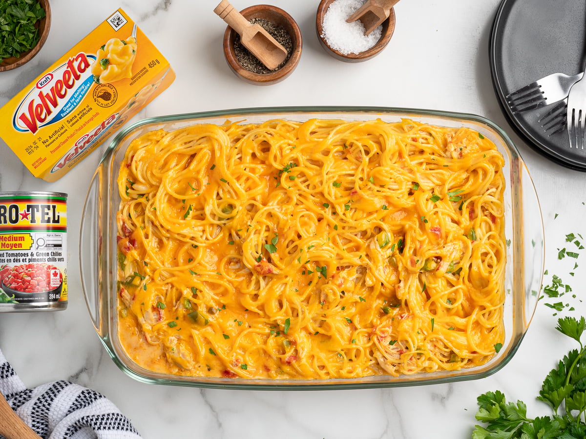A glass baking dish filled with creamy Rotel Chicken Spaghetti, topped with chopped herbs, sits on a marble counter. Nearby are a block of Velveeta cheese, a can of Ro-Tel, seasonings, and utensils.