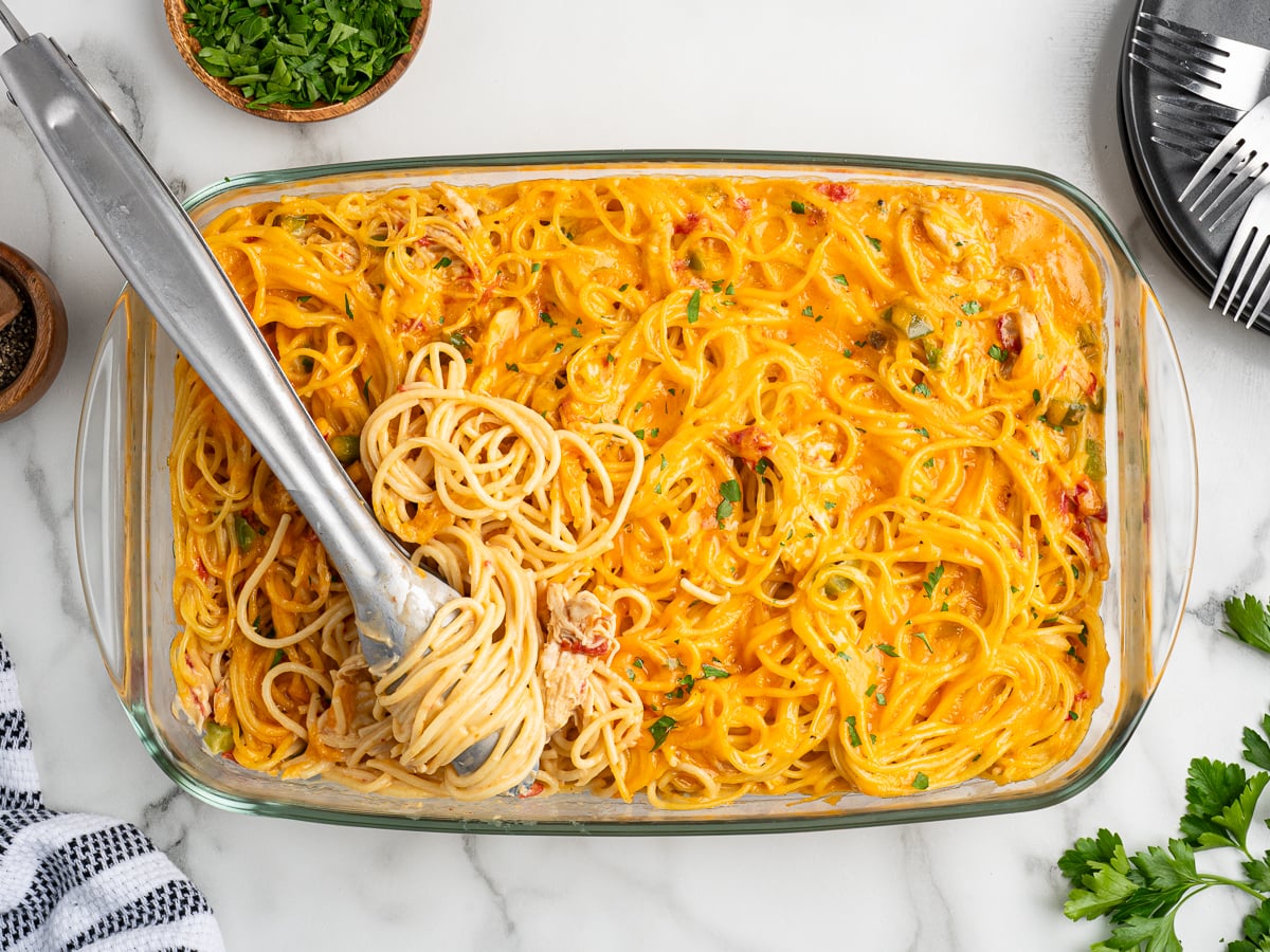 A glass baking dish filled with creamy Rotel Chicken Spaghetti, mixed with sauce and herbs. Metal tongs are lifting a serving of spaghetti. Plates, parsley, and a napkin are nearby on a marble surface.