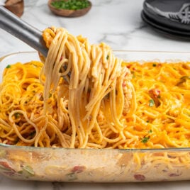 A glass baking dish filled with cheesy Rotel Chicken Spaghetti, with tongs lifting a serving. The creamy pasta is mixed with vegetables and chopped herbs, while plates and utensils rest on a marble countertop in the background.