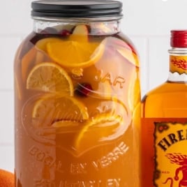 Tall dispenser filled with Fireball apple cider sangria packed with oranges, apples, and cranberries beside a Fireball whisky bottle.