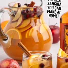 Apple cider sangria with Fireball whisky served in glasses and a pitcher with sliced apples, oranges, and fresh cranberries.