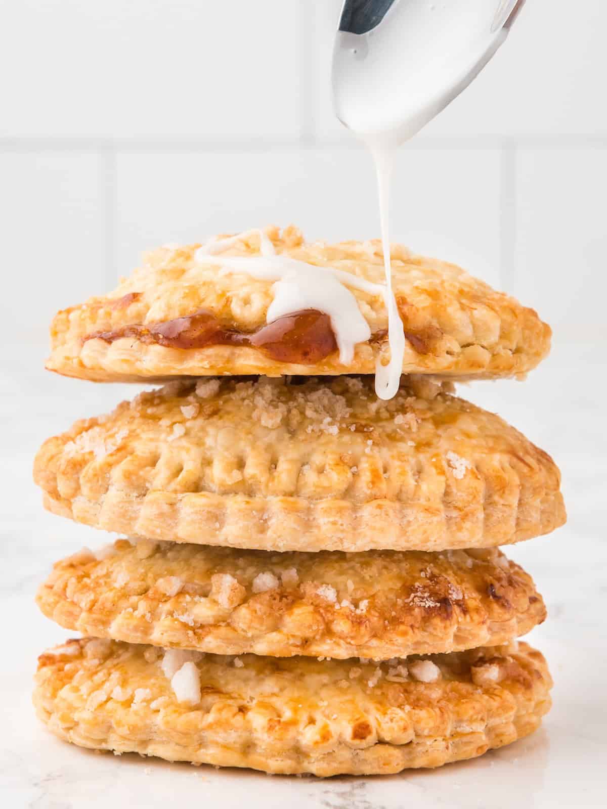 A stack of four round Air Fryer Hand Pies is shown, with icing being drizzled from a spoon onto the top pie. The pies have a golden, flaky crust and some visible sugar crystals.