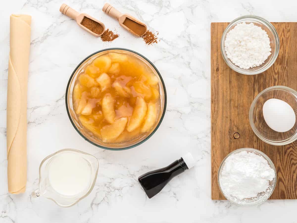 Bowl of apple pie filling, pie crust, milk, vanilla, egg, sugar, flour, cinnamon, and nutmeg are arranged on a marble surface and wooden board—perfectly prepped for making delicious Air Fryer Hand Pies.