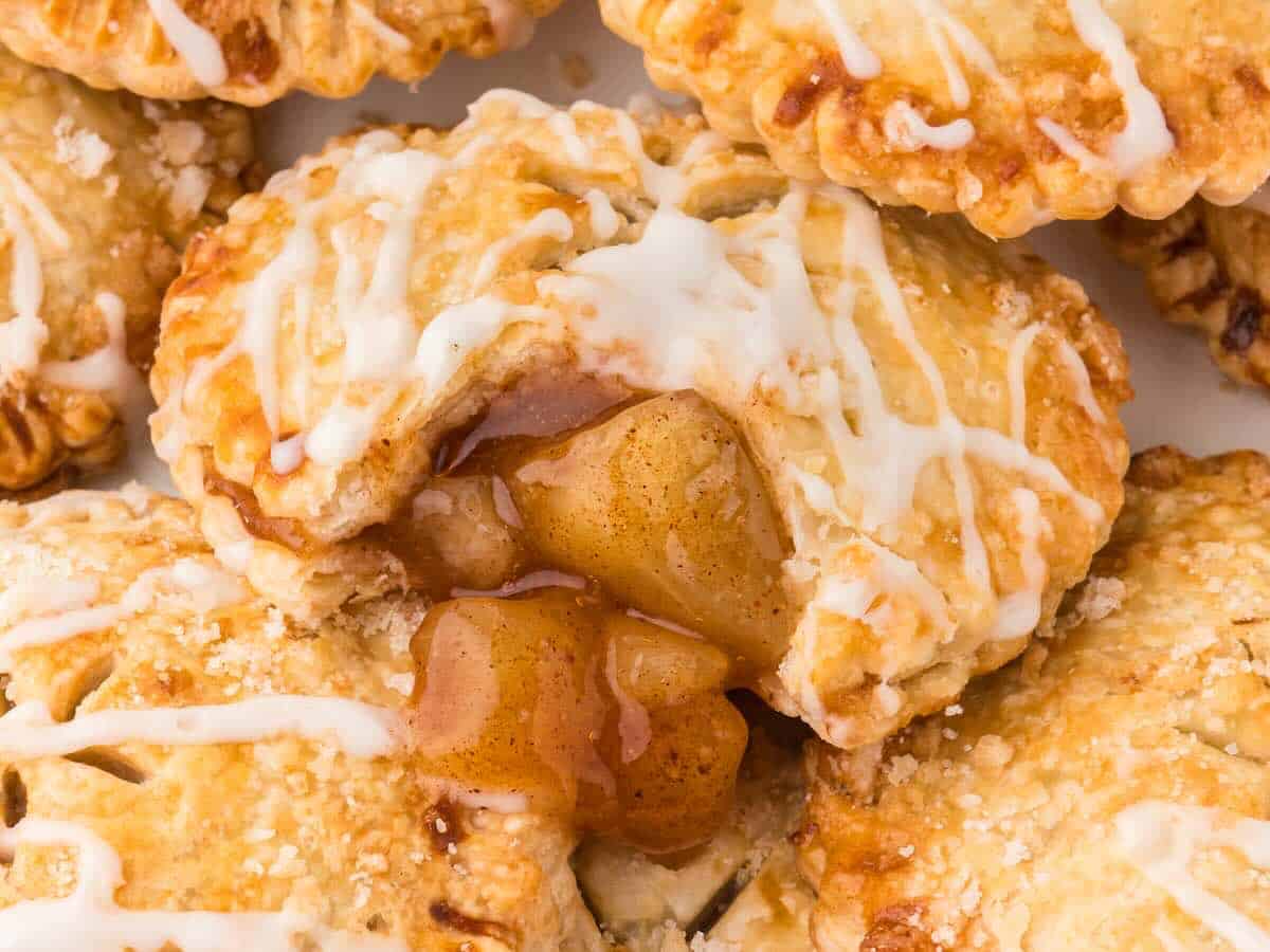 Close-up of a golden, flaky Air Fryer Hand Pie filled with gooey cinnamon apple filling, drizzled with white icing and sprinkled with coarse sugar. The pie is partially open, revealing the warm, spiced apple mixture inside.