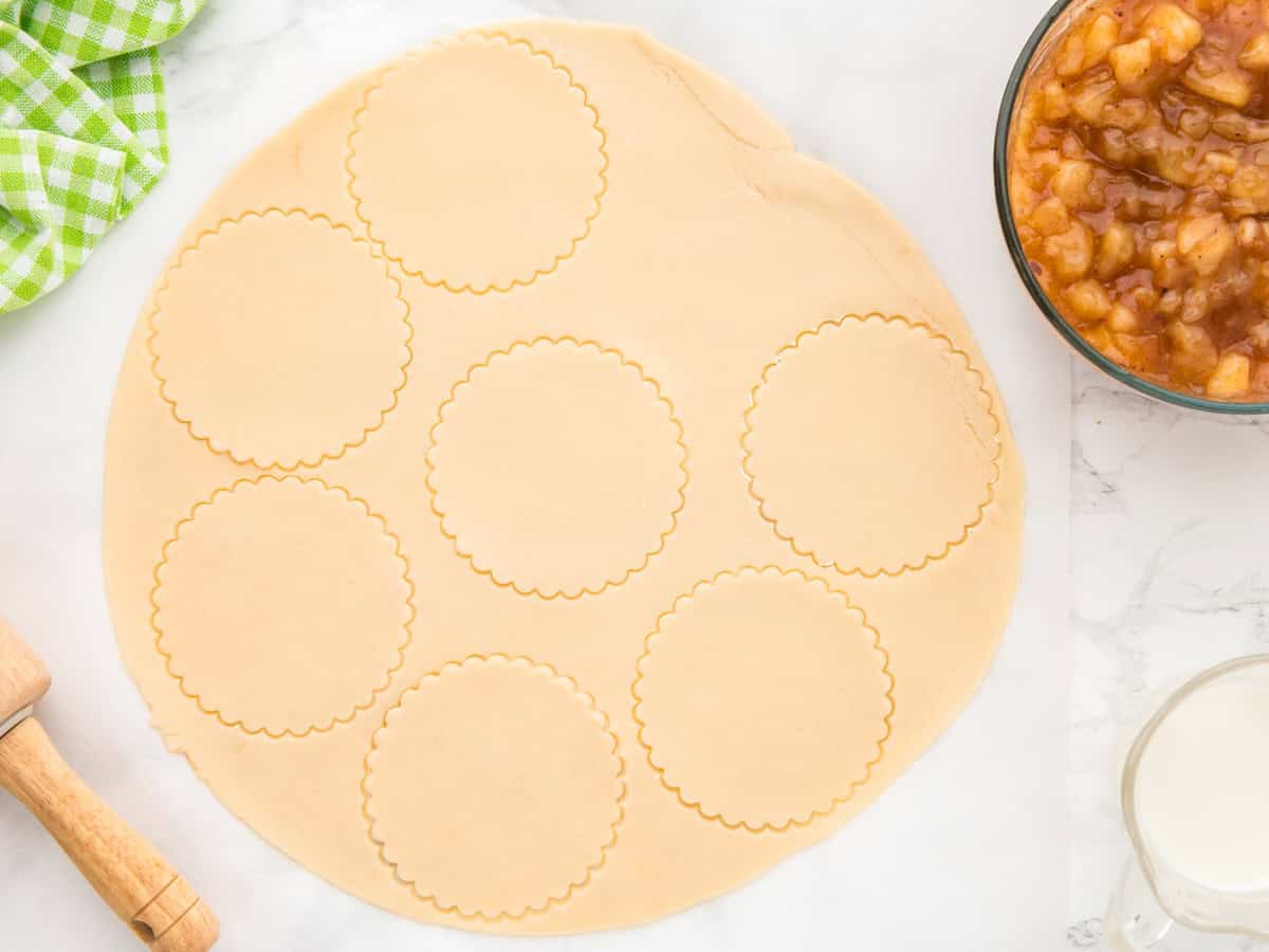 Round scalloped shapes are cut into rolled-out dough, ready to be transformed into delicious Air Fryer Hand Pies, with a rolling pin, a bowl of apple filling, a glass of milk, and a green checkered cloth nearby on a white surface.