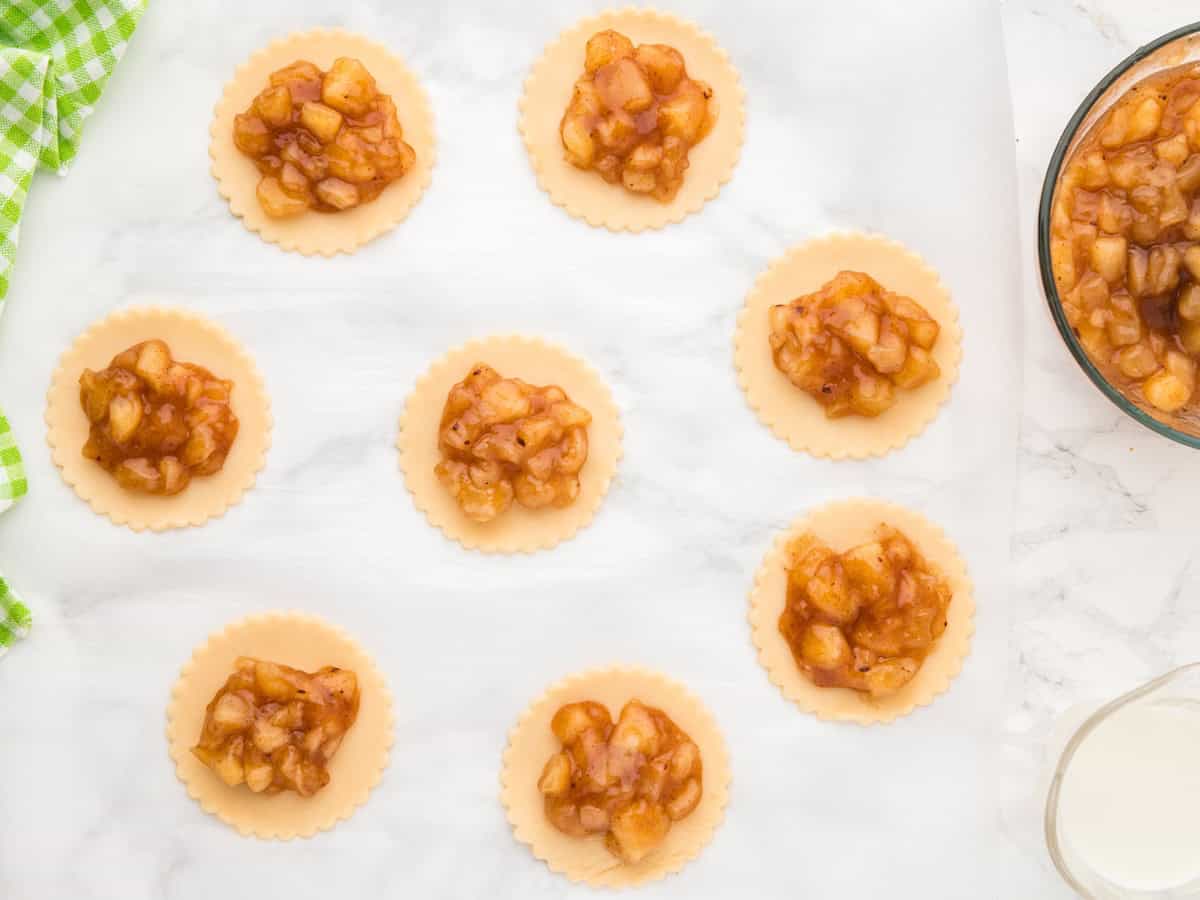 Eight round pastry dough circles topped with chunky apple pie filling are arranged on parchment paper, ready for baking as delicious Air Fryer Hand Pies. A bowl of extra apple filling is on the side. The background is a marble countertop.