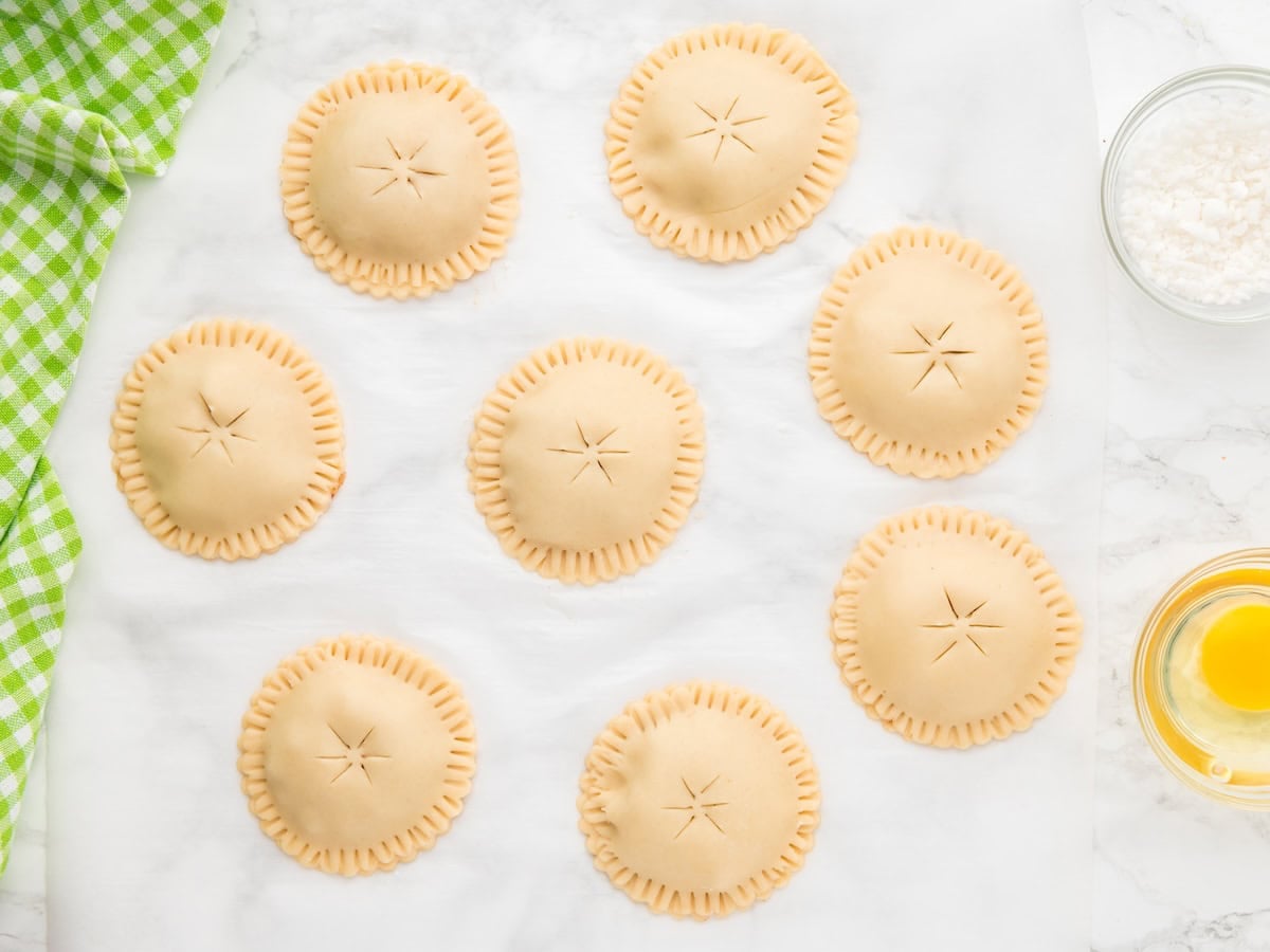 Eight unbaked round Air Fryer Hand Pies with crimped edges and star-shaped slits sit on parchment paper. A green checkered cloth, a bowl of sugar, and a bowl with an egg are nearby.