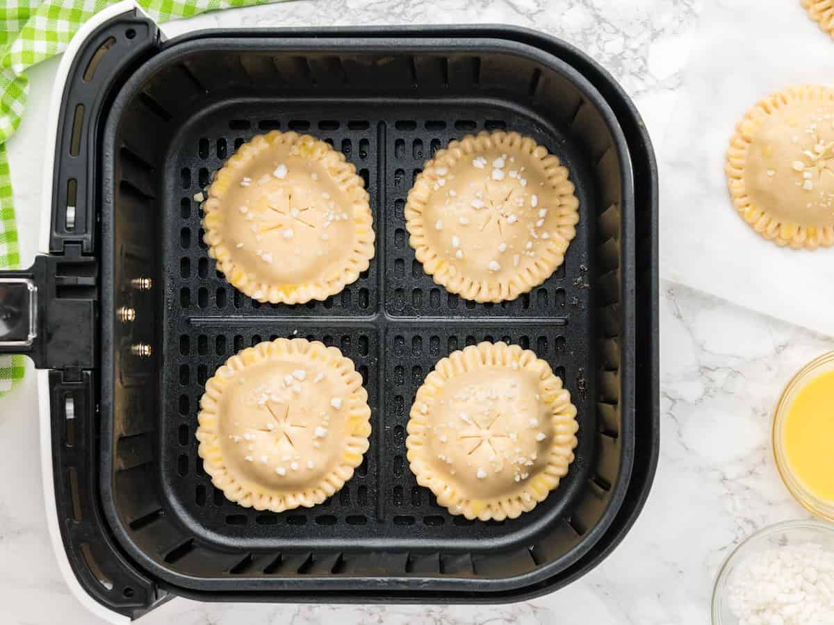 Four uncooked round Air Fryer Hand Pies with crimped edges and coarse sugar on top are arranged in a black air fryer basket, ready to be baked. A napkin, egg wash, and more hand pies are nearby on a marble counter.