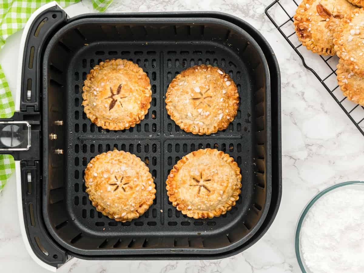 Four golden-brown Air Fryer Hand Pies are arranged in an air fryer basket. A cooling rack with more pies and a bowl of sugar are nearby on a marble countertop.