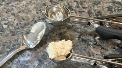 A metal spoon and two small metal scoops with black handles rest on a speckled countertop. One scoop contains a beige dough-like mixture for Chicken and Dumplings; the spoon also has a bit of the mixture on it, ready to be added to your favorite Chicken and Dumplings Recipe.