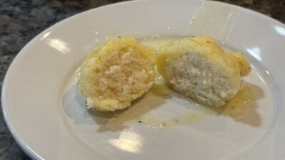 A dumpling cut in half sits on a white plate, covered in a light, translucent sauce—reminiscent of classic Chicken and Dumplings. The inside of the dumpling appears soft and fluffy. The plate rests on a dark, speckled countertop.