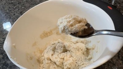 A spoon holds up a portion of sticky, unbaked dough from a large white mixing bowl on a granite countertop—just the beginning steps in preparing a comforting Chicken and Dumplings recipe.