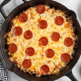 Overhead view of cast iron skillet with baked crustless pepperoni pizza surrounded by plates, forks, and extra cheese.