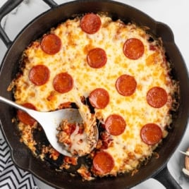 Cast iron skillet filled with cheesy pepperoni-topped crustless pizza, with a metal spoon serving a slice from the center.