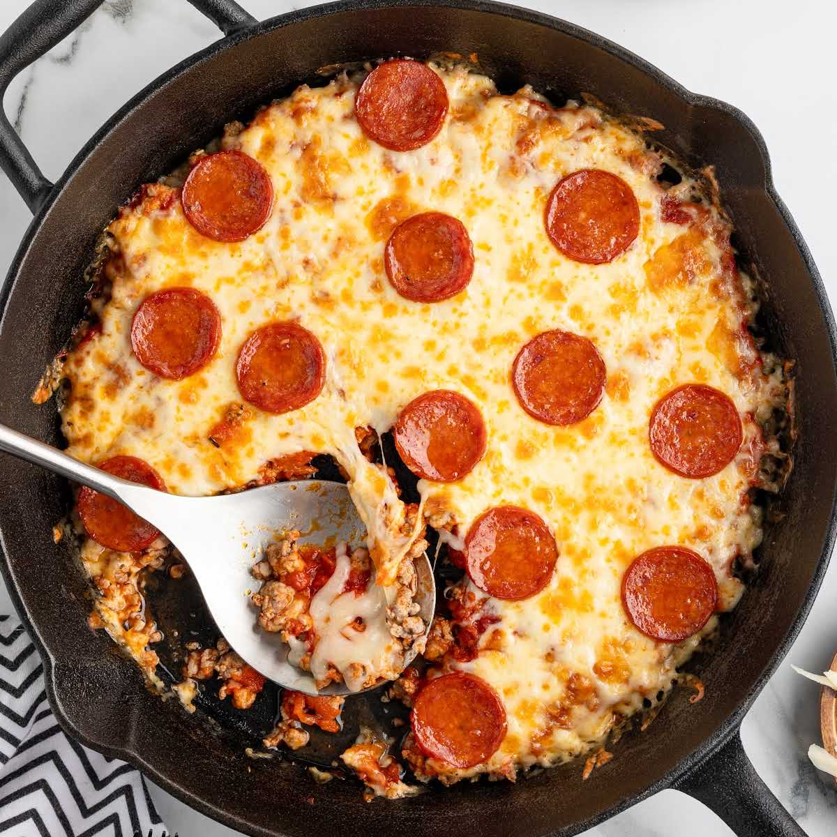 Cast iron skillet filled with cheesy pepperoni-topped crustless pizza, with a metal spoon serving a slice from the center.