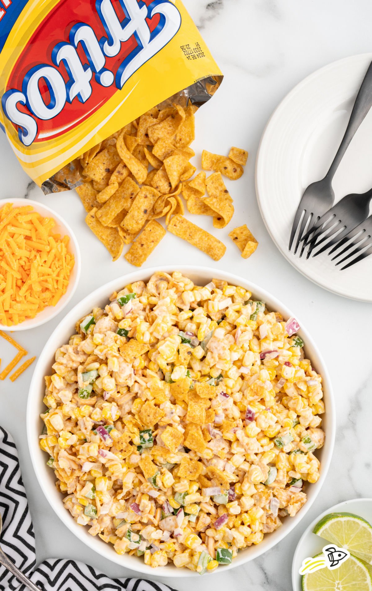 Overhead view of Fritos corn salad in a white bowl, topped with crushed Fritos, surrounded by shredded cheddar, a bag of Fritos, forks, and lime wedges on a marble background.
