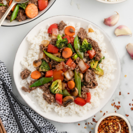 A white plate with a serving of ground beef stir fry on a bed of white rice.