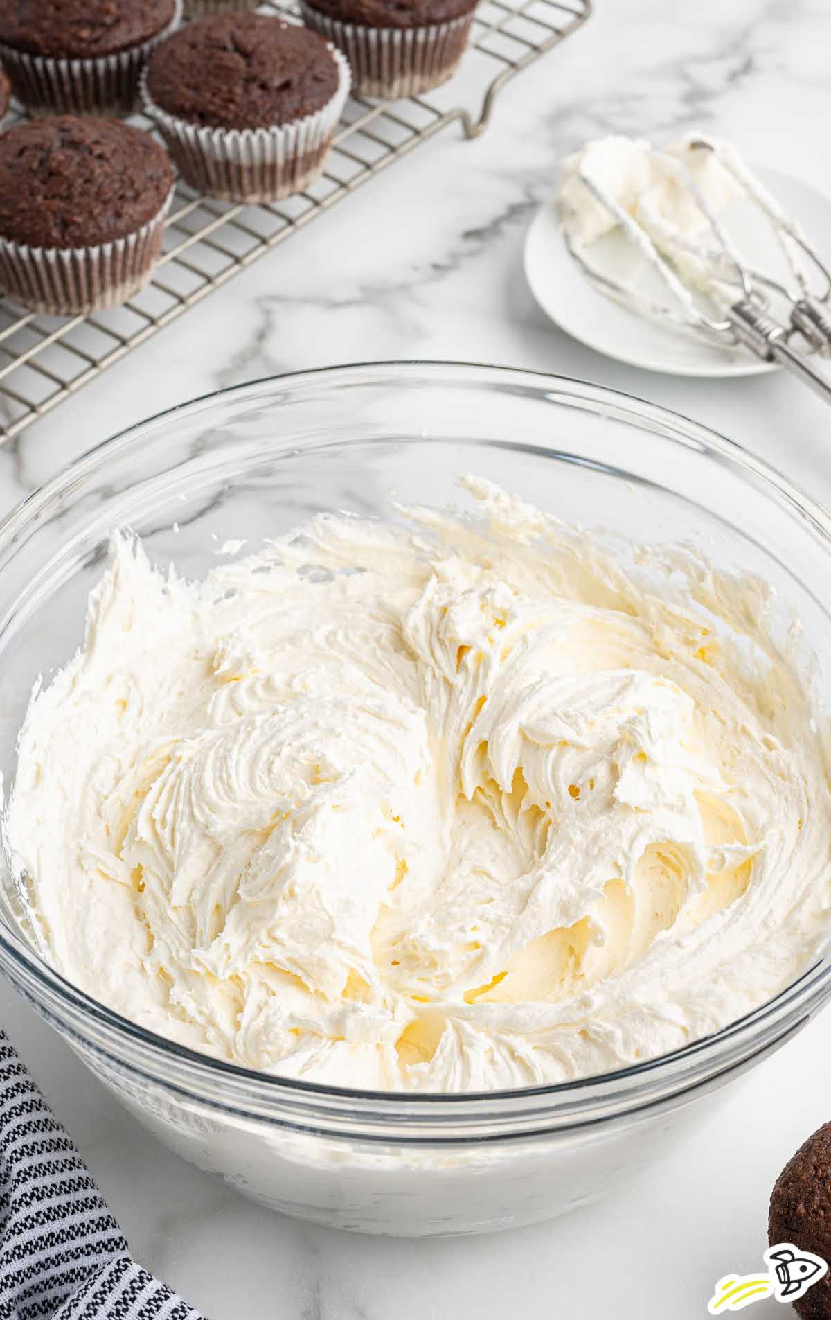 Large bowl of fluffy marshmallow frosting with chocolate cupcakes cooling on rack in background
