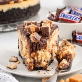 a slice of Snickers Cheesecake topped with crushed Snickers on a plate