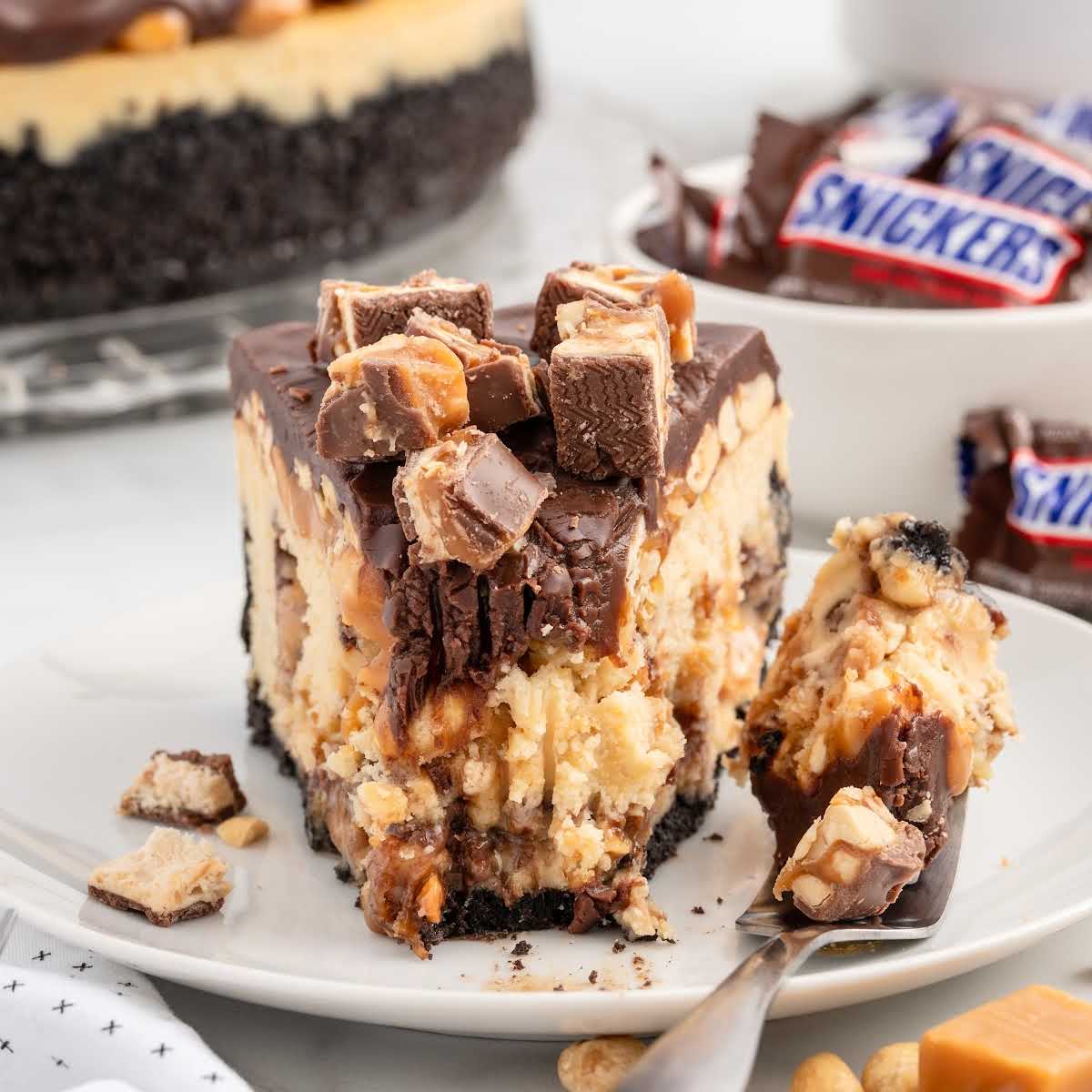 a slice of Snickers Cheesecake topped with crushed Snickers on a plate