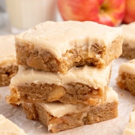 Stacked apple brownies with creamy frosting in front of fresh apples and a glass of milk