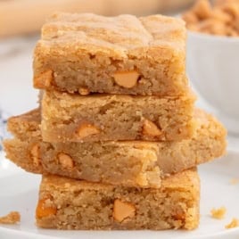 A stack of four golden brown Butterscotch Brownies with visible butterscotch chips sits on a white plate, with a bowl of butterscotch chips in the background.