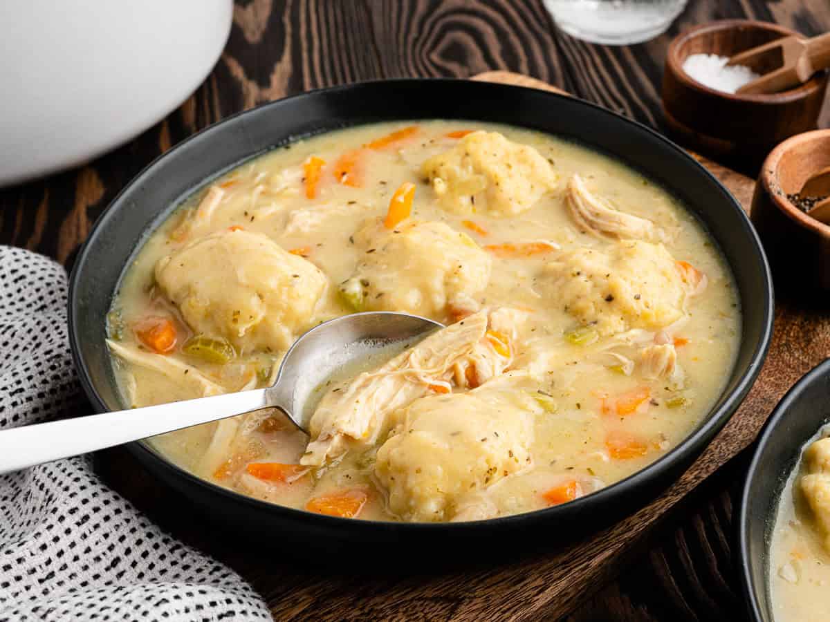 A bowl of Chicken and Dumplings features tender chunks of chicken, fluffy dumplings, carrots, and a savory broth. A spoon lifts chicken from the bowl, inviting you to enjoy this comforting Chicken and Dumplings Recipe. Nearby, a napkin and wooden spice bowls complete the cozy setting.