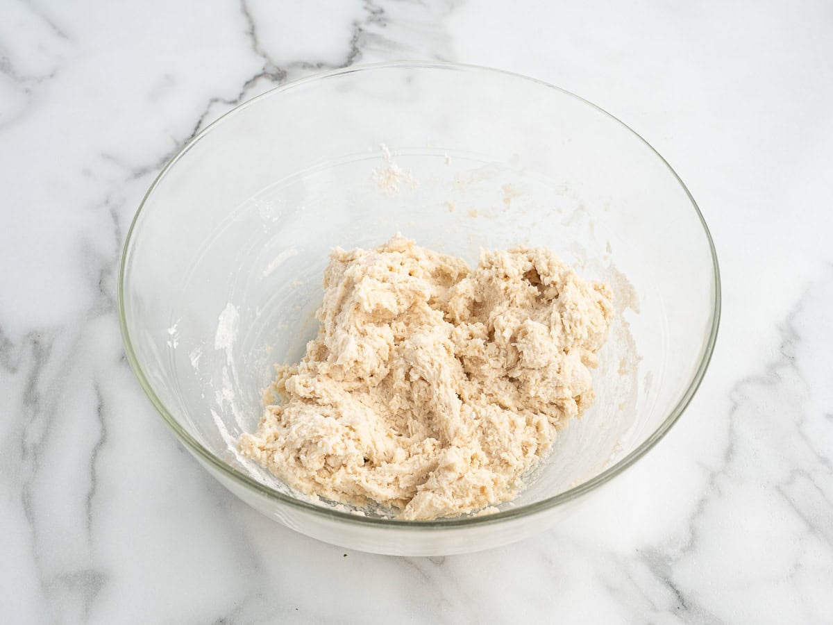 A glass bowl with rough, partially mixed dough sits on a white marble surface, ready to be transformed into delicious dumplings for your favorite Chicken and Dumplings Recipe.