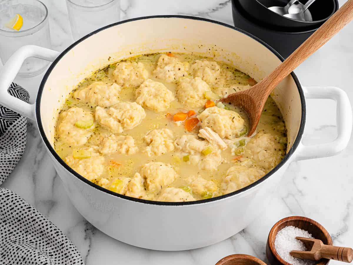 A white pot filled with a classic Chicken and Dumplings Recipe sits on a marble counter. A wooden spoon rests inside, and chopped vegetables and dumplings are visible in the savory broth.