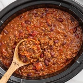 Overhead view of crockpot full of chili with a wooden spoon inside, surrounded by shredded cheese, chopped green onions, and a checkered towel.