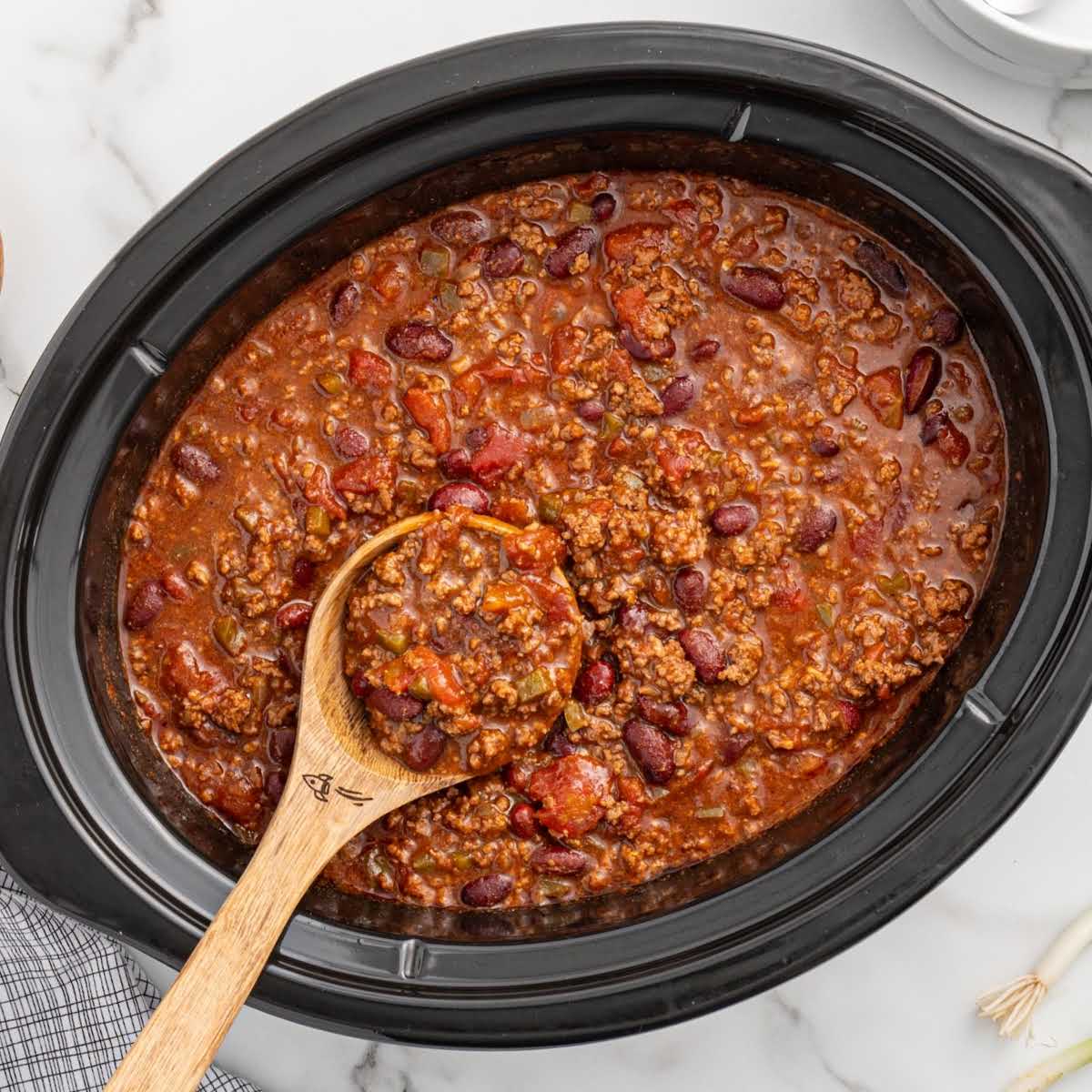 Overhead view of crockpot filled with chili and a wooden spoon scooping some out, with a cloth napkin nearby.