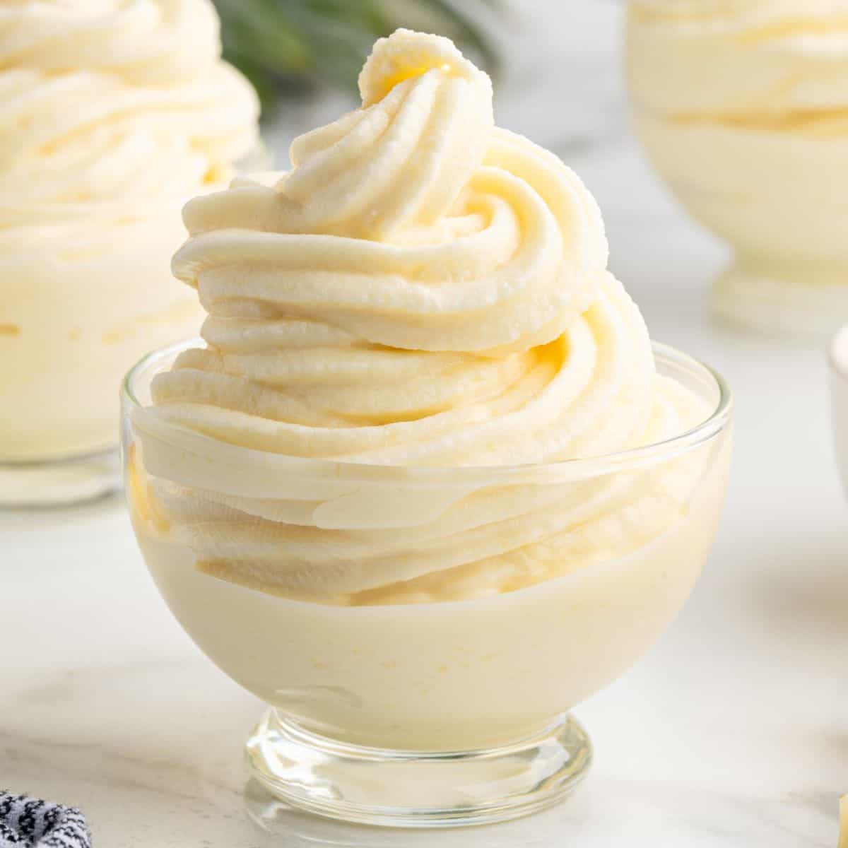 A glass dish filled with creamy, swirled pineapple soft serve, made from a classic Dole Whip Recipe, sits on a white surface with more dessert cups visible in the background.