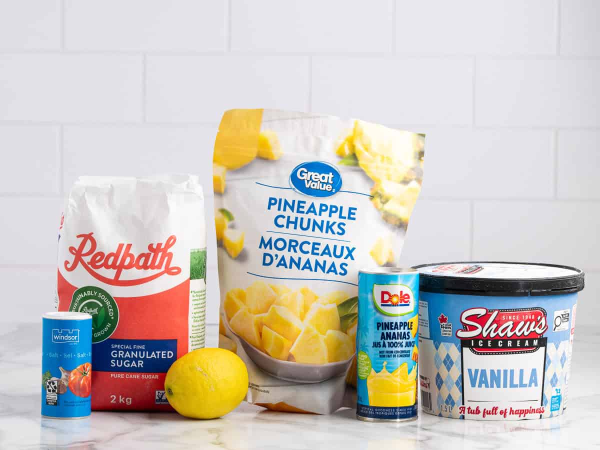 Assorted ingredients on a white counter: salt, granulated sugar, a lemon, pineapple chunks, canned pineapple, and a tub of vanilla ice creamโeverything you need for a homemade Dole Whip recipeโset against a white tile background.