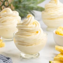 Three glass bowls of swirled pineapple soft serve inspired by the classic Dole Whip recipe, garnished with fresh pineapple slices and a whole pineapple in the background on a white surface.