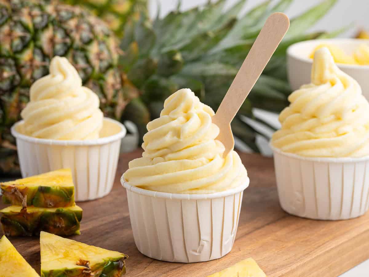 Three cups of creamy pineapple soft serve ice cream, inspired by the classic Dole Whip recipe, sit on a wooden surface surrounded by fresh pineapple slices and a whole pineapple in the background. One cup has a wooden spoon sticking out, ready to enjoy this delightful Dole Whip treat.
