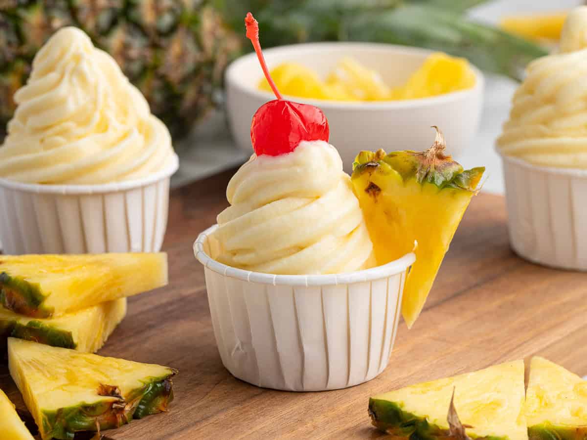 A swirl of pineapple Dole Whip soft serve in a white cup, topped with a cherry and a pineapple slice, sits on a wooden surface with pineapple pieces around it.