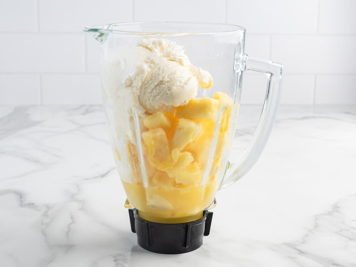 A blender filled with scoops of vanilla ice cream and orange juice sits on a marble countertop with a white tile background, ready to be blended into a creamy treat reminiscent of a classic Dole Whip recipe.