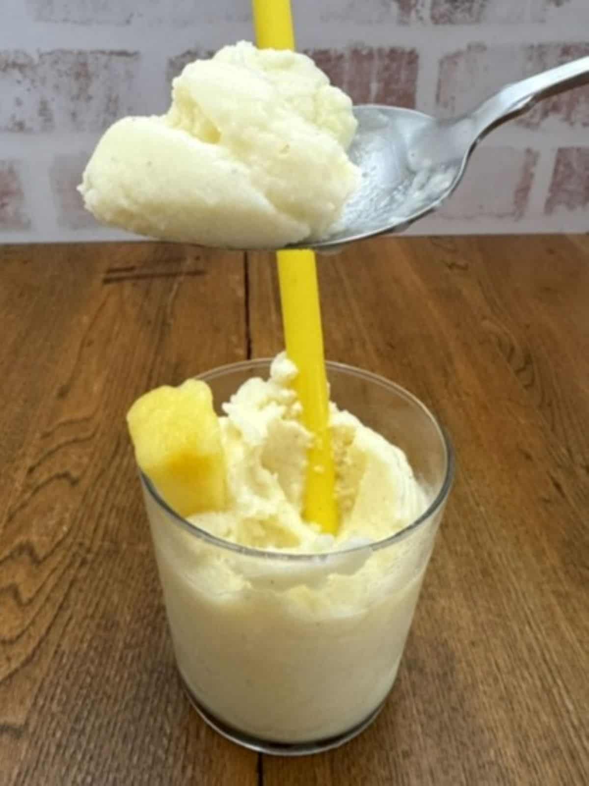A glass filled with creamy pale yellow Dole Whip ice cream, featuring a yellow straw and a frozen pineapple chunk, sits on a wooden table. A spoon holds a scoop of the Dole Whip above the glass.