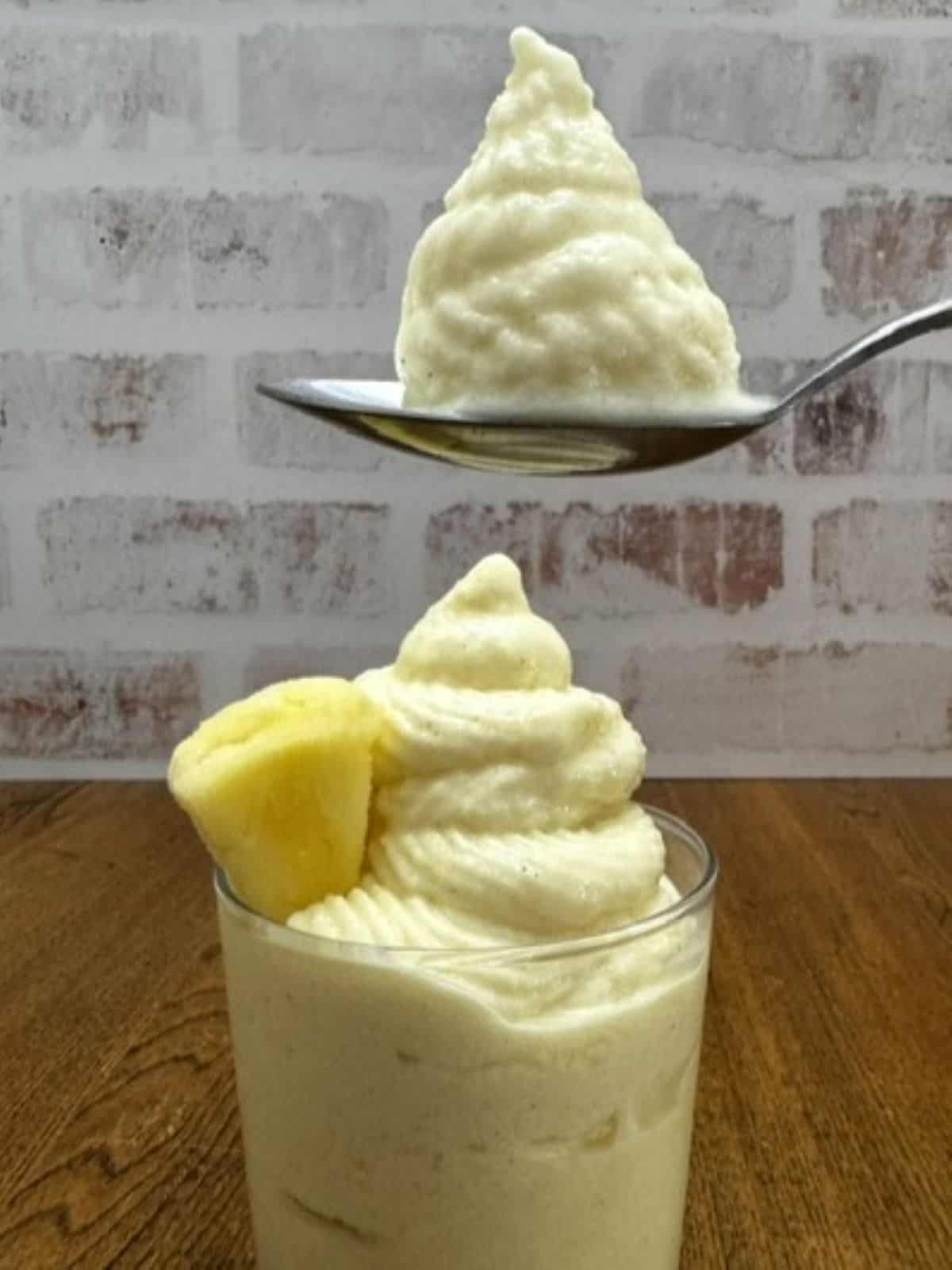 A glass filled with creamy, swirled banana soft serve inspired by the classic Dole Whip recipe sits on a wooden surface, while a spoon holding a scoop of the same luscious dessert is positioned above the glass.