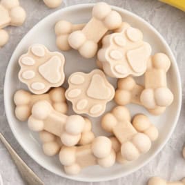 A white plate filled with creamy, bone-shaped and paw print-shaped frozen dog treats sits on a light surface. A spoon lies nearby.